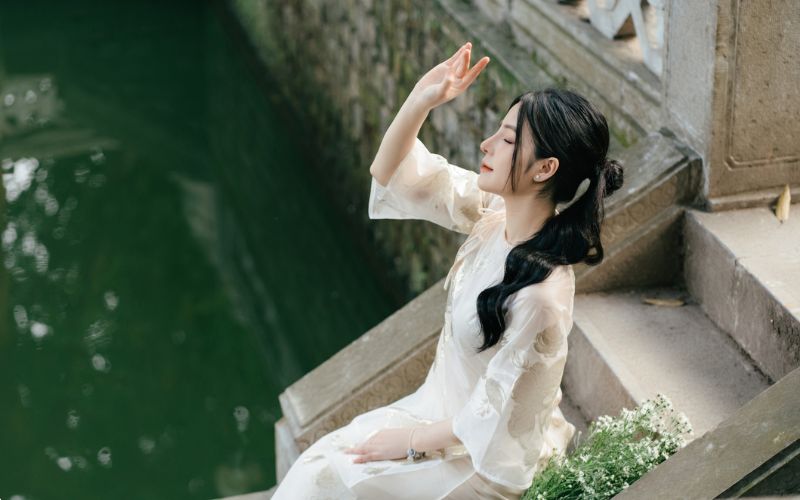 Young woman poses for Tet (Vietnamese New Year) photos in traditional ao dai dress, sitting gracefully by the lake.